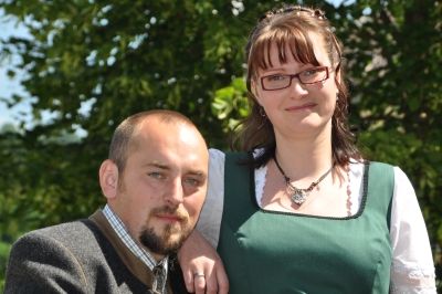 Ein Mann und eine Frau stehen eng beieinander, lächeln und posieren für ein Foto. Der Mann trägt eine graue Jacke und die Frau ein grünes Kleid und eine Brille. Sie stehen vor einem Baum.