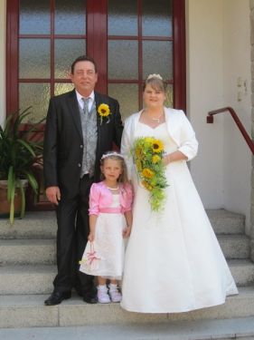 Ein Mann, eine Frau und ein junges Mädchen stehen lächelnd auf Stufen. Der Mann trägt einen schwarzen Anzug, die Frau ein weißes Hochzeitskleid und das Mädchen ein rosa Kleid. Sie halten einen Strauß Sonnenblumen.