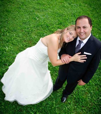 Ein frisch verheiratetes Paar, die Braut in einem weißen Kleid und der Bräutigam in einem schwarzen Anzug, posiert für ein Foto auf einem grünen Rasen, beide lächelnd und halten Händchen.