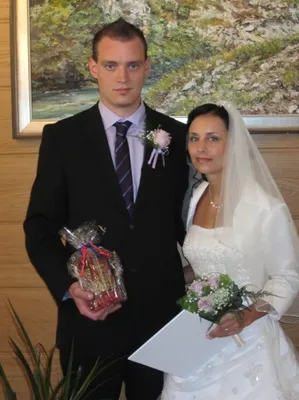 Ein Mann und eine Frau stehen zusammen, der Mann hält ein Geschenk und die Frau eine Blumenstrauß. Sie sind in formeller Kleidung gekleidet, wahrscheinlich für eine Hochzeit.
