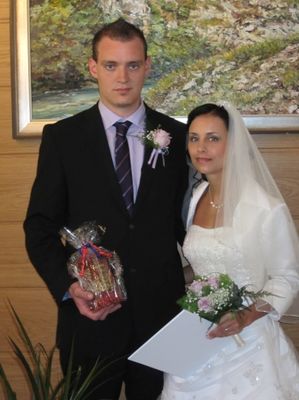 Ein Mann und eine Frau stehen zusammen, der Mann hält ein Geschenk und die Frau eine Blumenstrauß. Sie sind in formeller Kleidung gekleidet, wahrscheinlich für eine Hochzeit.