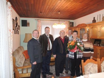 Eine Gruppe von Menschen, darunter zwei Frauen mit Blumen, stehen in einem Wohnzimmer mit Holzmöbeln und einer Holzdecke.