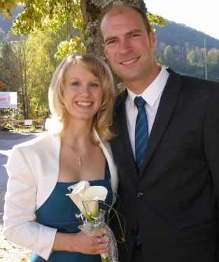 Ein Mann und eine Frau in formeller Kleidung stehen nah beieinander und lächeln, mit einem Berg und Bäumen im Hintergrund. Die Frau hält einen Strauß weißer Blumen.