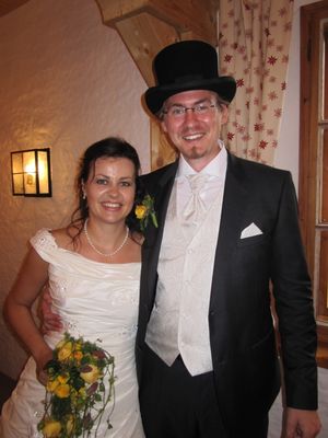 Ein Mann und eine Frau in formeller Kleidung, lächeln für ein Foto. Die Frau trägt ein weißes Hochzeitskleid und hält einen Blumenstrauß. Der Mann trägt einen schwarzen Anzug mit weißem Weste und einem schwarzen Zylinder.