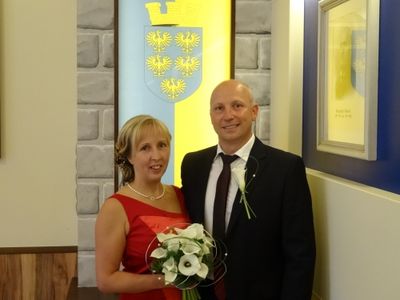 Ein Mann und eine Frau stehen vor einer Wand mit einem Wappen. Die Frau hält einen Blumenstrauß.