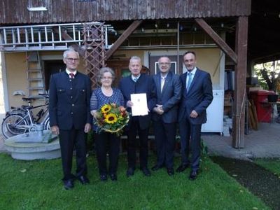 Fünf Personen stehen auf einem Rasen vor einem Haus, einer hält einen Blumenstrauß und ein anderer ein Zertifikat. Ein Fahrrad und eine Leiter sind im Hintergrund zu sehen.