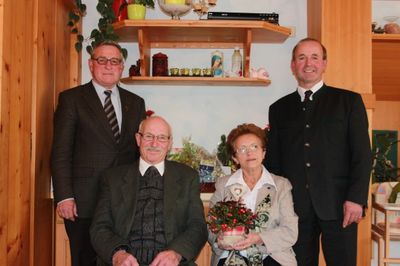 Vier Personen stehen in einem Raum mit Holzwänden, zwei Männer in Anzügen, ein älterer Mann in einem Pullover und eine Frau mit Blumen. Dahinter befindet sich ein Regal mit verschiedenen Gegenständen.