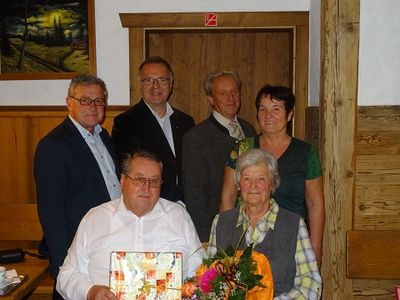 Eine Gruppe von Menschen steht in einem Raum, lächelt und posiert für ein Foto. Ein Mann vorne hält eine Geschenkbox und einen Blumenstrauß. Hinter ihm sitzt eine Frau und hält einen Blumenstrauß. Ein Mann steht hinter ihnen, und eine Frau steht neben ihm. Ein Bilderrahmen ist an der Wand montiert.