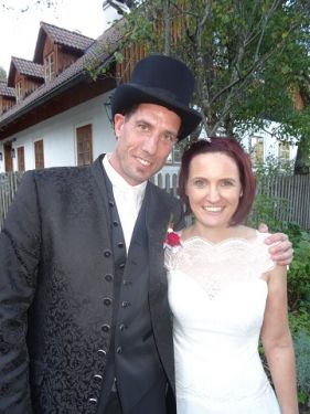Ein Mann in einem schwarzen Zylinderhut und eine Frau in einem weißen Hochzeitskleid stehen nebeneinander vor einem Haus.