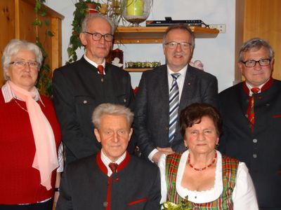 Eine Gruppe älterer Menschen in traditioneller Kleidung posiert für ein Foto in einem Raum mit Holzregalen und Pflanzen im Hintergrund.