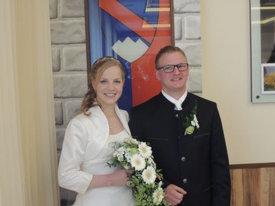 Ein frisch verheiratetes Paar steht lächelnd nebeneinander, die Braut in einem weißen Kleid und der Bräutigam in einem schwarzen Anzug, vor einer dekorativen Wand.
