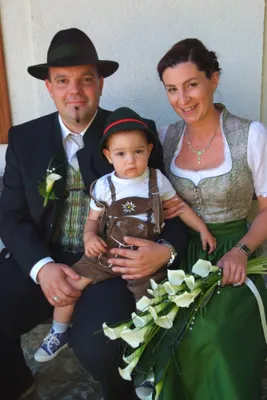 Eine Familie zu dritt, gekleidet in traditioneller deutscher Kleidung, lächelt und hält einen Blumenstrauß. Der Mann trägt einen Hut, die Frau eine Halskette und das Baby eine traditionelle Kleidung.