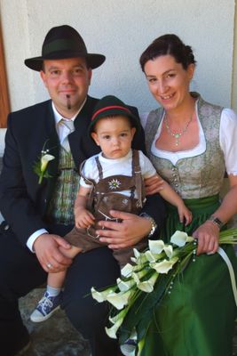 Eine Familie zu dritt, gekleidet in traditioneller deutscher Kleidung, lächelt und hält einen Blumenstrauß. Der Mann trägt einen Hut, die Frau eine Halskette und das Baby eine traditionelle Kleidung.