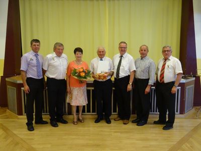 Eine Gruppe von sechs Personen, darunter Männer und Frauen, stehen zusammen und lächeln für ein Foto. Der Mann in der Mitte hält einen Blumenstrauß und ein Buch.