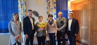 Fünf Personen stehen zusammen in einem Raum und lächeln für ein Foto. Die Frau in der Mitte hält einen Blumenstrauß. Hinter ihnen befindet sich eine Wand mit einem dekorativen Blumenstück und blauen Vorhängen.