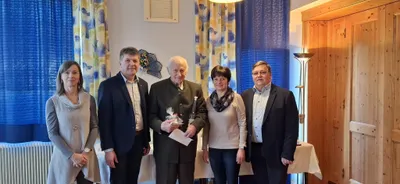 Vier Menschen stehen zusammen in einem Raum und lächeln für ein Foto. Der Mann in der Mitte hält eine Flasche und ein Papier. Die Frau neben ihm trägt eine Schürze. Der Mann rechts trägt eine Brille.
