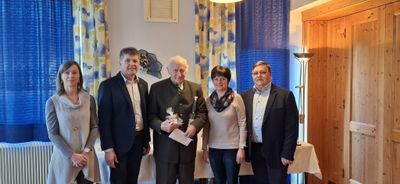 Vier Menschen stehen zusammen in einem Raum und lächeln für ein Foto. Der Mann in der Mitte hält eine Flasche und ein Papier. Die Frau neben ihm trägt eine Schürze. Der Mann rechts trägt eine Brille.