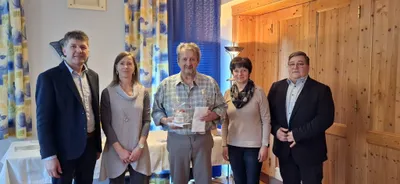 Ein Mann, der ein Geschenk und ein Papier in der Hand hält, steht zwischen zwei Frauen und einem weiteren Mann in einem Raum mit Holzwänden und Vorhängen.