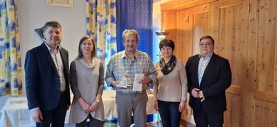 Ein Mann, der ein Geschenk und ein Papier in der Hand hält, steht zwischen zwei Frauen und einem weiteren Mann in einem Raum mit Holzwänden und Vorhängen.
