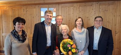 Vier Personen stehen lächelnd in einem Raum für ein Foto. Eine ältere Frau hält einen Blumenstrauß. Hinter ihr stehen ein Mann mit Brille und eine Frau in einem grauen Pullover neben einem Mann im Anzug.