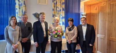 Vier Personen stehen in einem Raum mit einem Tisch und lächeln. Eine Frau hält einen Blumenstrauß. Hinter ihnen befinden sich blaue Vorhänge und ein dekoratives Wandstück.