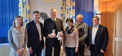Fünf Personen stehen lächelnd für ein Foto zusammen. Ein Mann in der Mitte hält ein verpacktes Geschenk und Papier. Dahinter sind eine Blumendekoration und ein Vorhang zu sehen.