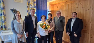 Vier Menschen stehen lächelnd in einem Raum. Eine Frau hält einen Blumenstrauß. Hinter ihnen befindet sich eine Holzwand und ein blauer Vorhang.