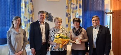 Drei Menschen stehen lächelnd in einem Raum. Eine Frau hält einen Strauß gelber Blumen. Eine andere Frau hält ein verpacktes Geschenk. Ein Mann steht zwischen ihnen.
