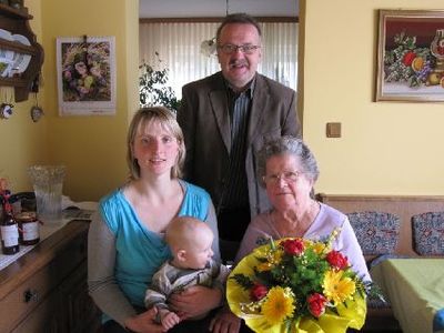 Ein Familienfoto mit einer Frau, die ein Baby hält, einem Mann, der hinter ihnen steht, und einer älteren Frau, die mit einem Blumenstrauß sitzt. Der Hintergrund zeigt gelbe Wände und gerahmte Bilder.