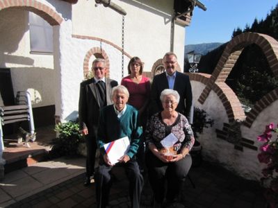 Fünf Personen stehen vor einem Haus, zwei sitzend, halten Papiere und eine Schüssel, mit einem Ziegelbogen und Berg im Hintergrund.