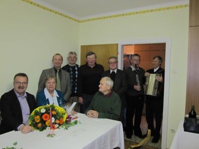 Eine Gruppe von Menschen, einschließlich eines älteren Mannes, versammelt sich um einen Tisch mit einem Blumenarrangement. Einige halten Musikinstrumente.