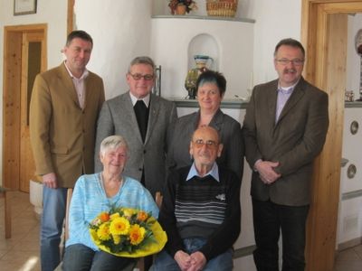 Eine Gruppe von sechs Personen, einschließlich eines älteren Paares, posiert für ein Foto in einem Raum mit weißen Wänden und Holzakzenten. Der ältere Mann sitzt auf einem Stuhl, und die ältere Frau hält einen Strauß gelber Blumen.
