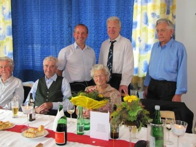 Eine Gruppe älterer Menschen, darunter eine Frau mit einem Blumenstrauß, sitzt an einem Tisch mit Weingläsern, Flaschen und Essen und lächelt für ein Foto.