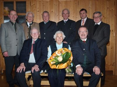 Eine Gruppe von Männern und einer älteren Frau posiert für ein Foto vor einer Holzwand. Die ältere Frau hält einen Blumenstrauß.