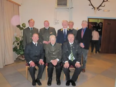Eine Gruppe von Männern in formeller Kleidung, möglicherweise ein Verein oder eine Organisation, posiert für ein Foto in einem Raum mit einer weißen Wand und einem aufgehängten Hirschkopf.