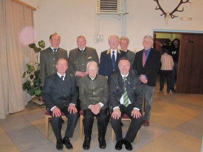 Eine Gruppe von Männern in formeller Kleidung, möglicherweise ein Verein oder eine Organisation, posiert für ein Foto in einem Raum mit einer weißen Wand und einem aufgehängten Hirschkopf.