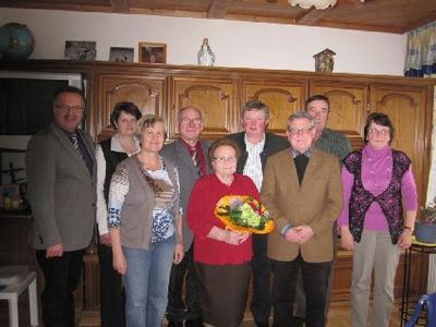 Eine Gruppe von Menschen in formeller Kleidung, die in einem Raum stehen, lächeln und für ein Foto posieren. Eine Frau in der Mitte hält einen Blumenstrauß. Hinter ihnen sind Holzschränke und ein Fernseher zu sehen.