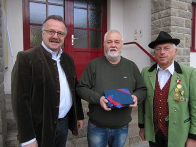 Drei Männer stehen vor einem Gebäude, einer hält eine blaue Geschenkbox. Der Mann links trägt eine Brille und einen braunen Mantel, der mittlere Mann einen grünen Pullover und der Mann rechts einen grünen Mantel mit Medaillen.