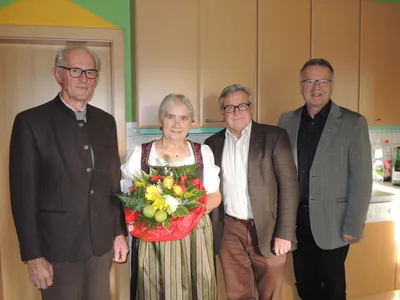 Vier Personen stehen in einem Raum, eine Frau hält einen Blumenstrauß. Die Frau trägt ein traditionelles Kleid. Hinter ihr stehen drei Männer in formeller Kleidung. Im Hintergrund befinden sich Holzschränke und eine grüne Wand.