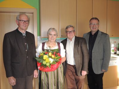 Vier Personen stehen in einem Raum, eine Frau hält einen Blumenstrauß. Die Frau trägt ein traditionelles Kleid. Hinter ihr stehen drei Männer in formeller Kleidung. Im Hintergrund befinden sich Holzschränke und eine grüne Wand.