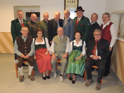 Eine Gruppe älterer Männer und Frauen in traditioneller Kleidung, die in Stühlen sitzen, möglicherweise für ein Gruppenfoto. Sie lächeln und schauen in die Kamera.