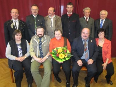 Eine Gruppe von Männern und Frauen, alle in formeller Kleidung, posiert für ein Foto vor einem roten Vorhang. Eine Person hält einen Blumenstrauß.