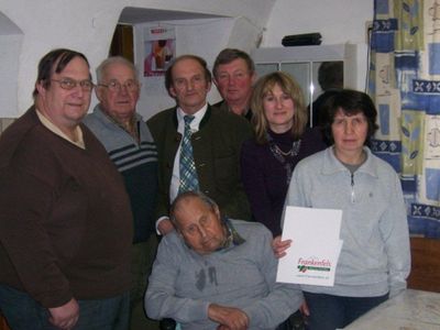 Eine Gruppe von Menschen, einschließlich eines älteren Mannes im Rollstuhl, versammelt sich in einem Raum. Eine Frau in einem grauen Pullover hält ein Zertifikat. Der Raum hat weiße Wände und einen Vorhang mit Blattdesign.