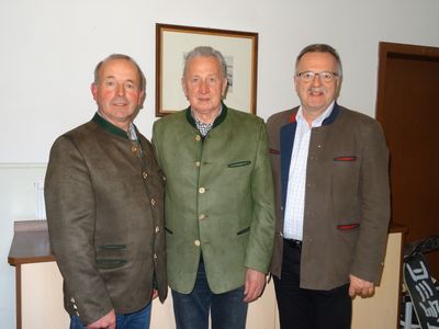 Drei Männer in traditioneller Kleidung stehen zusammen vor einer Wand mit einem gerahmten Bild dahinter.