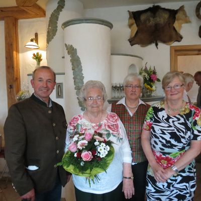 Eine Gruppe von vier Personen, die zusammenstehen und für ein Foto lächeln. Eine ältere Frau hält einen Blumenstrauß, und ein Mann steht zu ihrer Linken. Dahinter sind eine weiße Säule und ein Pelzfell zu sehen.