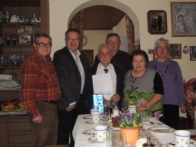 Eine Gruppe älterer Menschen versammelt sich um einen Esstisch, lächelt und posiert für ein Foto. Der Tisch ist mit Tellern, Tassen, Weingläsern und einer kleinen Topfpflanze gedeckt. Ein Mann in der Mitte hält ein Zertifikat.