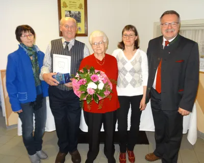 Fünf Menschen stehen lächelnd für ein Foto zusammen. Eine ältere Frau hält einen Blumenstrauß. Ein Mann hält ein Zertifikat und eine Geschenkbox. Hinter ihnen ist ein gerahmtes Bild an der Wand angebracht.