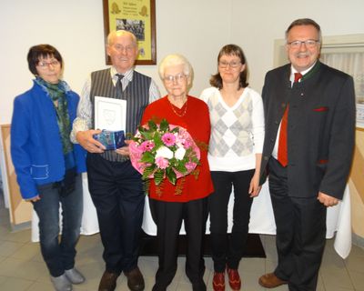 Fünf Menschen stehen lächelnd für ein Foto zusammen. Eine ältere Frau hält einen Blumenstrauß. Ein Mann hält ein Zertifikat und eine Geschenkbox. Hinter ihnen ist ein gerahmtes Bild an der Wand angebracht.