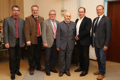 Fünf Männer in formeller Kleidung stehen zusammen in einem Raum und lächeln für ein Foto. Der Mann in der Mitte trägt einen grauen Anzug.