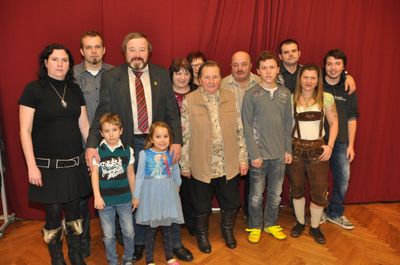 Eine Gruppe von Menschen, darunter Erwachsene und Kinder, posiert für ein Foto vor einem roten Vorhang. Der Mann in der Mitte trägt eine Krawatte und eine Anstecknadel.